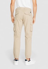 Beige Cargo-Hosen mit elastischen Bündchen, zwei Seitentaschen und zwei Gesäßtaschen. Kombiniert mit weißen Sneakers mit marineblauen Akzenten.