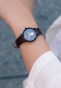 Montre noire ronde avec un cadran bleu foncé, des index horaires blancs et un petit indicateur de date, montée sur un bracelet en cuir marron.