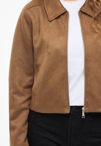 Osoba oblečená v hnedej semišovej bunde na zips cez biele tričko a čierne nohavice, zobrazená od ramien po pás na bielom pozadí.