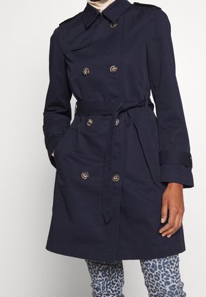 Laivastonsininen kaksirivinen trenssitakki, jossa vyö ja napitetut hihansuut, käytetty sinisten ja harmaiden leopardikuvioisten housujen päällä.