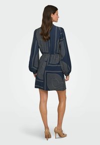Marineblauwe jurk met witte liniaire patronen, lange pofferige mouwen, een gathered taille en een korte zoom. G gedragen met naakte hoge hakken.