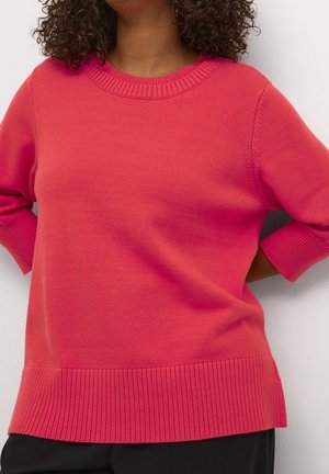 Kobieta o kręconych włosach ubrana w jasnoróżowy dziany sweter z prążkowanymi mankietami, krawędzią i dekoltem, stojąca na tle białego tła.