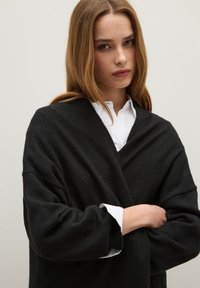 Cardigan nero oversize in maglia con maniche larghe, indossato sopra una camicia bianca con colletto. Texture morbida, design minimale e scollatura a V.