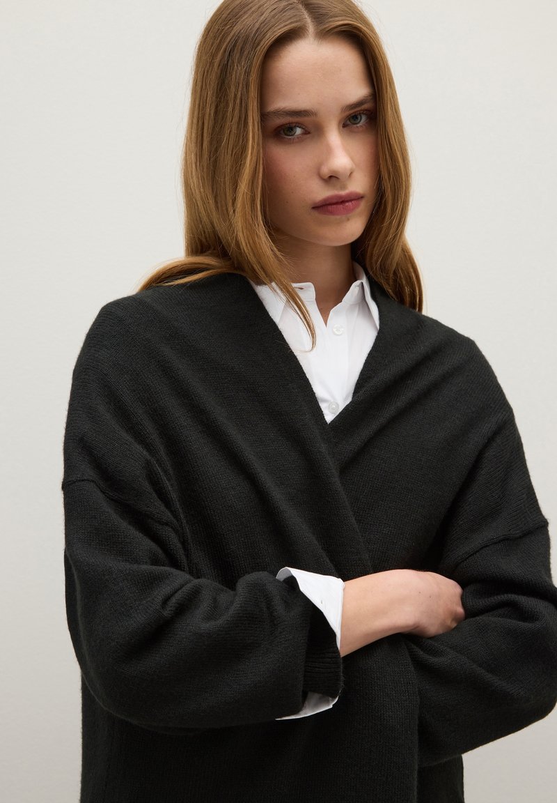 Cardigan nero oversize in maglia con maniche larghe, indossato sopra una camicia bianca con colletto. Texture morbida, design minimale e scollatura a V.