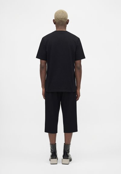 Άνδρας με κοντά ξανθά μαλλιά, φοράει μαύρο κοντομάνικο πουκάμισο, μαύρα βερμούδα μήκους μέχρι το γόνατο και μαύρα μποτάκια με ασημί τετράγωνα τακούνια, γυρισμένος από πίσω.