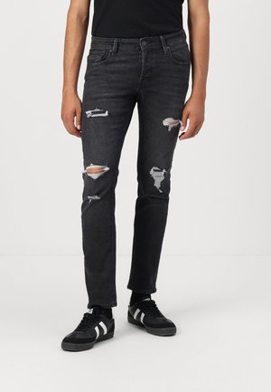 Mann trägt eng anliegende schwarze Jeans mit zerrissenen Knien und schwarze Sneakers mit weißen Streifen, steht vor einem schlichten Hintergrund.