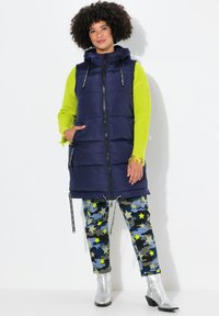 Marinblå puffer-väst med huva, dragkedja och sidofickor. Kombinerad med kamoflagobyxor med stjärnmönster och blanka silverfärgade ankelstövlar.