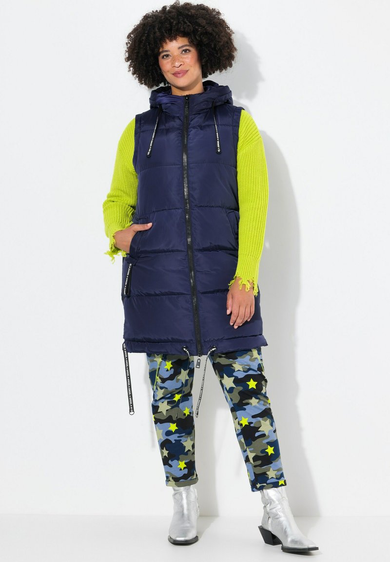 Marinblå puffer-väst med huva, dragkedja och sidofickor. Kombinerad med kamoflagobyxor med stjärnmönster och blanka silverfärgade ankelstövlar.