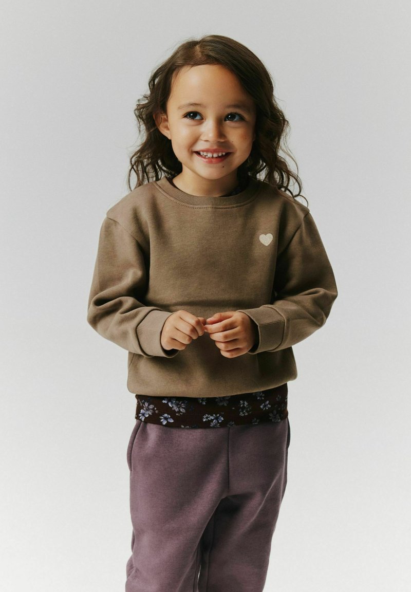 Jeune enfant souriant, portant un sweat-shirt marron avec un petit cœur et un pantalon violet, debout devant un fond clair uni.