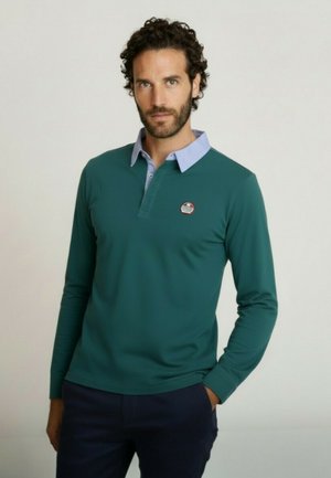 Homme aux cheveux bouclés portant un polo à manches longues bleu sarcelle avec un col bleu clair et un pantalon bleu marine, debout devant un fond uni.