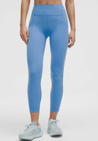 Lichtblauwe high-waisted leggings met een gladde textuur, twee zijzakken en een aansluitend ontwerp. Gecombineerd met lichtgrijze sportschoenen.