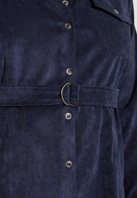 Mörkblå kordkjolsklänning med bälte i midjan, som har metallknappar och en bröstficka. Mjuk textur och strukturerad design.