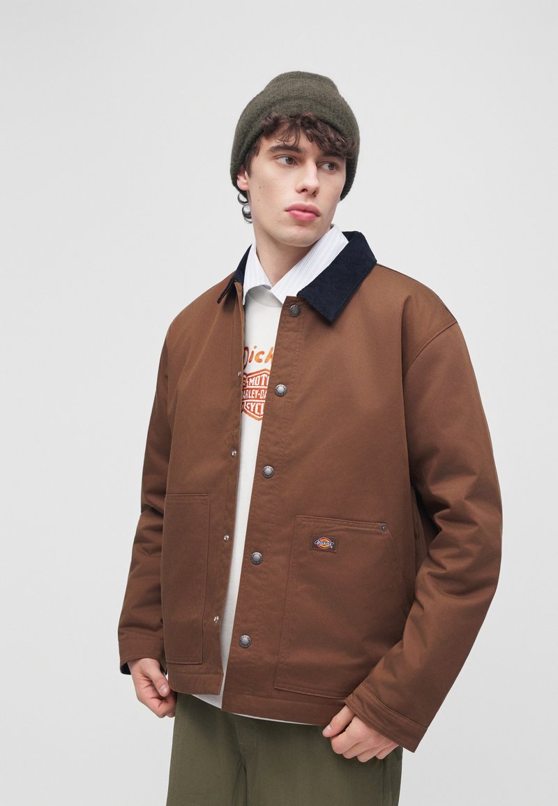 Young man wearing a brown buttoned jacket with large front pockets, a green beanie, white shirt, and printed T-shirt, looking left.