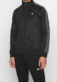 Chaqueta deportiva negra con cremallera y alto cuello, mangas largas con franjas laterales en blanco y azul, y un pequeño logo en el pecho.