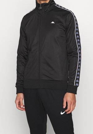 Veste de sport zippée noire avec col montant, manches longues arborant des bandes latérales blanches et bleues, et un petit logo sur la poitrine.