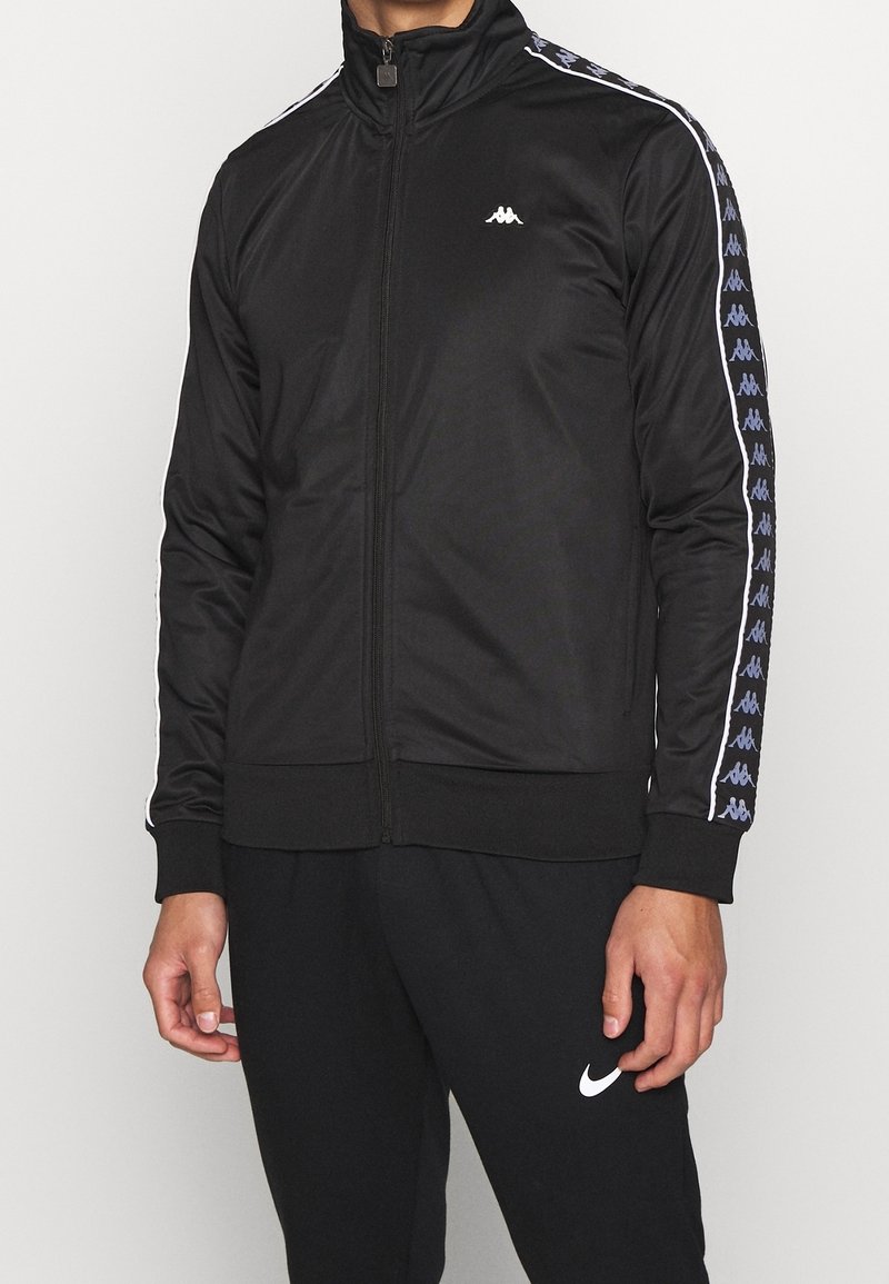 Chaqueta deportiva negra con cremallera y alto cuello, mangas largas con franjas laterales en blanco y azul, y un pequeño logo en el pecho.