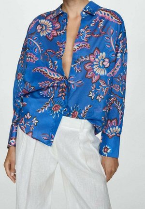 Blauwe blouse met lange mouwen en roze en paarse bloemenpatronen, deels opengeknipt, gecombineerd met witte broek met hoge taille.