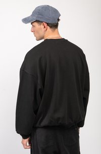 Sudadera negra de gran tamaño con un dobladillo acanalado, hecha de algodón. Combinada con una gorra azul deslavada, mostrando un ajuste relajado y hombros caídos.