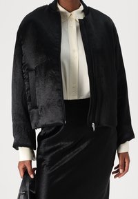 Svart bomberjacka med en mjuk, lätt krinklad textur, dragkedjestängning och sidofickor, bärs över en krämfärgad skjorta med knappar.