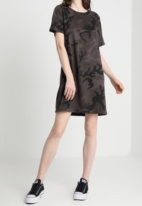 Short-sleeve, brown camouflage dress with a relaxed fit, made of soft fabric; paired with black sneakers featuring white soles.