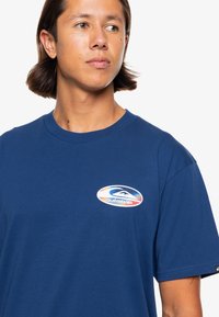 Navy blue cotton t-shirt featuring a colorful oval Quiksilver logo on the left chest and a classic crew neck design. Short sleeves.