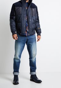 Marinblå pufferjacka med denimdetaljer, dragkedja framtill, ribbade ärmslut och flera fickor. Matchas med ljus tvättade jeans och marinblå stövlar.