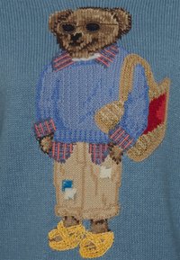 Sweter dziewiarski z haftowanym niedźwiedziem, który ma na sobie niebieski sweter, kraciastą wyściółkę, beżowe szorty, żółte kapcie i niesie torbę.