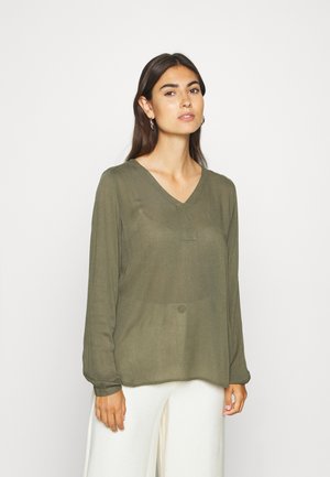 Olijfgroene blouse met lange mouwen en een V-hals, voorzien van knoopdetails aan de voorkant, gemaakt van transparant materiaal, gecombineerd met witte wijde broekspijpen.