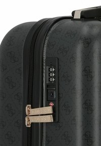 Esquina de maleta rígida negra con patrón de logo en relieve, tiradores de cremallera dorados de Guess y cerradura de combinación TSA ajustada a 000.