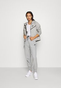 Graue Nike-Jacke mit Reißverschluss und schwarzen Akzenten, kombiniert mit grauen Jogginghosen und weißen Sneakers. Verfügt über eine Kapuze und einen verstellbaren Kordelzug in der Taille.