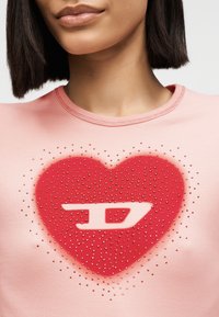 Γυναίκα που φοράει ροζ μπλούζα με κόκκινη καρδιά στολισμένη με μικρές στράς και ένα λευκό στυλιζαρισμένο "D" στο κέντρο.