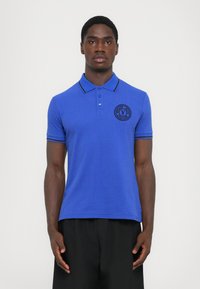 Βασιλικό μπλε πόλο μπλούζα με υφή, κουμπιά μπροστά και μαύρες ριγέ λεπτομέρειες στον γιακά και τα μανίκια. Εμφανές λογότυπο στο στήθος.