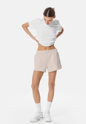 Kvinde iført hvid T-shirt, beige shorts, hvide sokker og hvide sneakers, der kigger ned, mens hun justerer sin skjorte.