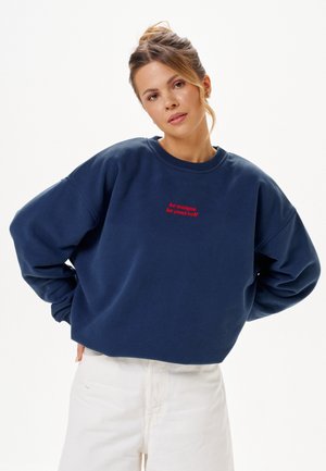 Femme aux cheveux attachés portant un sweat-shirt bleu marine avec un texte rouge « sois unique sois toi-même » et un pantalon blanc sur un fond uni.