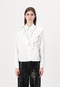 Camisa blanca con botones, con un frente fruncido y detalles de joyas, combinada con pantalones de lentejuelas negros. Tejido suave, diseño estructurado.
