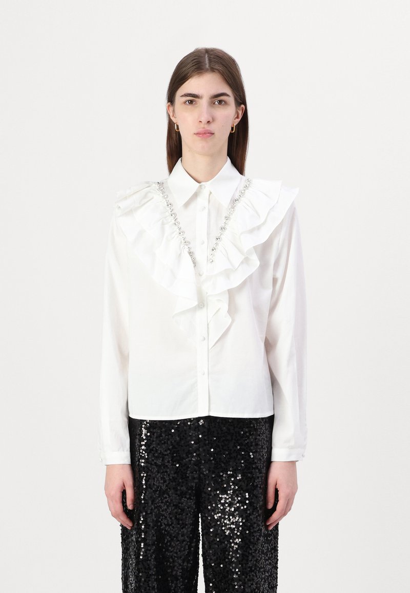 Camisa blanca con botones, con un frente fruncido y detalles de joyas, combinada con pantalones de lentejuelas negros. Tejido suave, diseño estructurado.