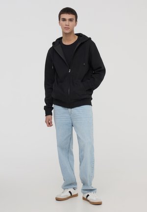 Czarna bluzka z kapturem na zamek błyskawiczny, noszona na czarnym t-shircie, w połączeniu z jasnoniebieskimi dżinsami i białymi trampkami z czarnymi akcentami.