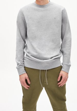 Mann trägt hellgraues Sweatshirt mit Rundhalsausschnitt und langen Ärmeln sowie olivgrüne Hose mit Kordelzug, steht vor einem einfarbigen weißen Hintergrund.