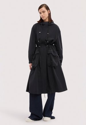 Kobieta ubrana w długi czarny płaszcz z kapturem i dużymi przednimi kieszeniami, szerokie ciemne dżinsy oraz białe trampki, stojąca na jednolitym tle.