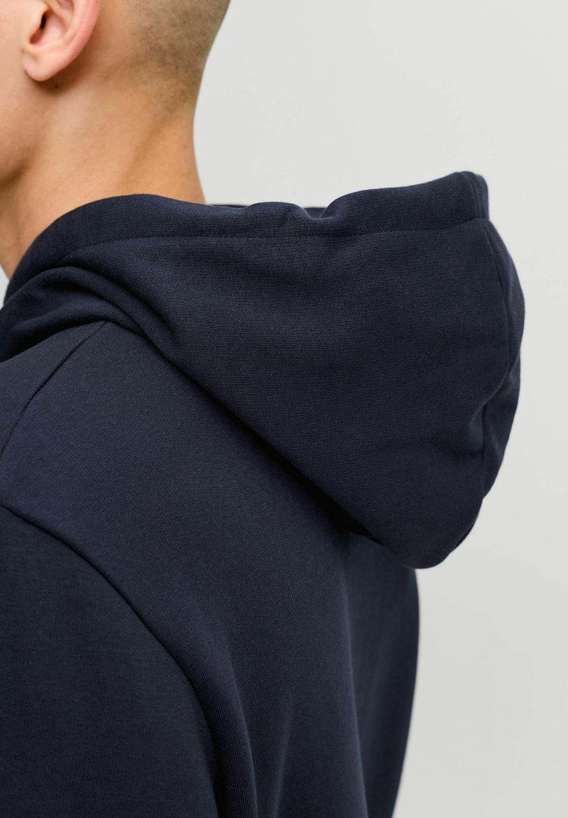 Personne aux cheveux courts portant un sweat à capuche bleu marine foncé, montrant l'arrière et le côté de la tête avec la capuche rabattue.
