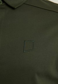 Temno zelena polo majica z mehkimi vlakni, z izrazitim kvadratnim logotipom izvezenim v svetlejšem odtenku na levi strani.