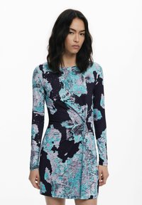 Frau mit schulterlangen dunklen Haaren, die ein langärmliges, knielanges Kleid mit blauem, schwarzem und grauem abstraktem Muster trägt, stehend vor einem schlichten weißen Hintergrund.