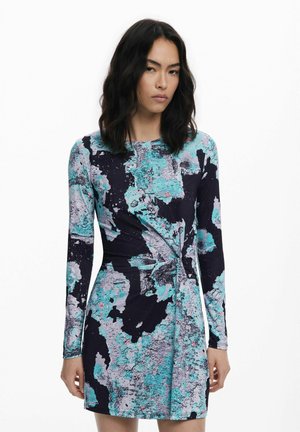Frau mit schulterlangen dunklen Haaren, die ein langärmliges, knielanges Kleid mit blauem, schwarzem und grauem abstraktem Muster trägt, stehend vor einem schlichten weißen Hintergrund.