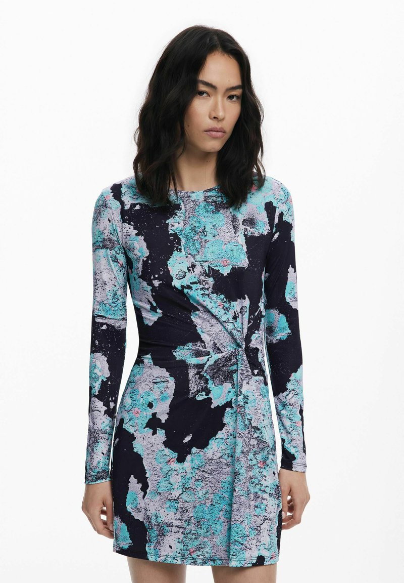 Frau mit schulterlangen dunklen Haaren, die ein langärmliges, knielanges Kleid mit blauem, schwarzem und grauem abstraktem Muster trägt, stehend vor einem schlichten weißen Hintergrund.
