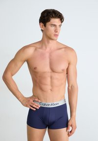 Marineblauwe strakke boxershorts met een grijze Calvin Klein tailleband. De stof is glad en rekbaar, wat een slanke lichaamsbouw accentueert.