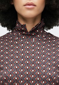 Blúzka s vysokým golierom v tmavosivej farbe s opakujúcim sa vzorom diamantov v bielej a červenej farbe. Hladká tkanina s jemným leskom a štruktúrovaným dizajnom.