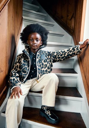 Dieťa s afro účesom oblečené v bunde a čižmách s leopardím vzorom, sediace na drevenom schodisku a držiace sa zábradlia jednou rukou.