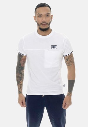 Uomo con capelli corti e barba indossa una camicia bianca a maniche corte con taschino sul petto e logo "LEONE 1947", pantaloni blu scuro, e ha tatuaggi su tutto il braccio.