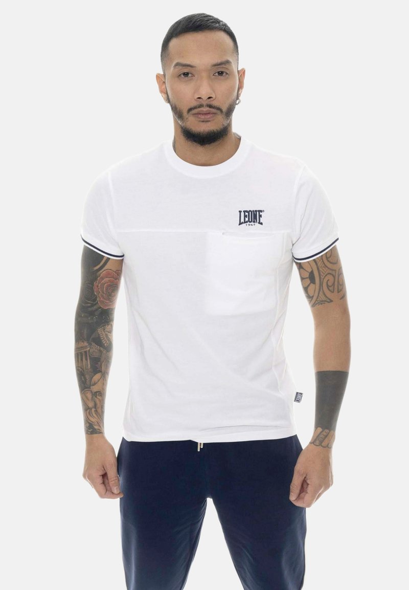 Homme aux cheveux courts et barbe porte une chemise blanche à manches courtes avec une poche poitrine et le logo "LEONE 1947", pantalon bleu marine, et a des tatouages sur tout le bras.