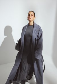 Naine kannab pikka halli trench coat'i tumeda halli kleidi üle, millel on frantsiääriga ääred, seistes ühetoonilise valge tausta ees.